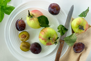 Several ripe plums, apples and a knife with a wooden handle lie on a white porcelain plate on a light painted wood surface. Horizontal orientation, top view, selective focus.