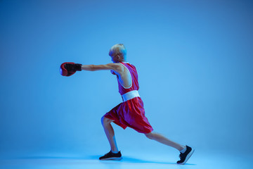 Fototapeta na wymiar Teenager in sportswear boxing isolated on blue studio background in neon light. Novice male caucasian boxer training hard and working out, kicking. Sport, healthy lifestyle, movement concept.