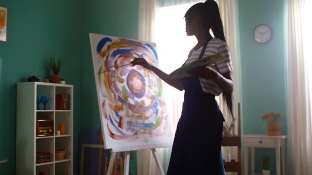 Young black woman is staying home, entertaining by painting on big canvas, using bright colors that attribute to her inner state, enjoys visual art, Zoom in, Slow motion.