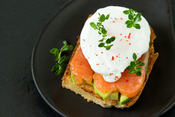 Poached egg with smoked salmon, toasted bread , avocado and coffee on on a black plate. Sandwich for breakfast.