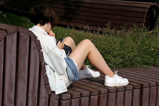 Slim girl in wireless headphones with naked legs in sneakers sitting with a smartphone on wooden bench on a city street. Concept of online addiction, sms, using phone outdoors
