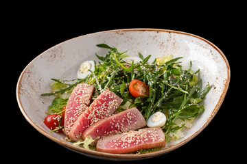 Salad with sliced steak of tuna in sesame, arugula, tomatoes and quail eggs. Tuna salad isolated on black.