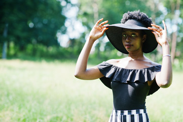 Fototapeta premium Portrait of gorgeous african american woman 20s in wear in black and white stripe pants and summer hat posing at green grass in park.