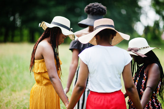 Group of four gorgeous african american womans wear summer hat holding hands and praying at green grass in park.