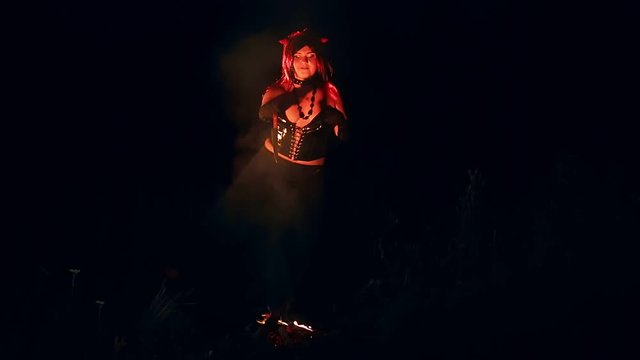 A woman in the image of a devil in black with horns at night by the fire with a knife dances a ritual dance.