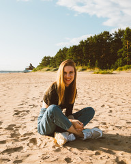 Full length beautiful smiling woman sitting on beach at sunset in evening. Pretty female with long brown hair, looking at camera. Slow life, social detox, self-immersion, being single concept. 