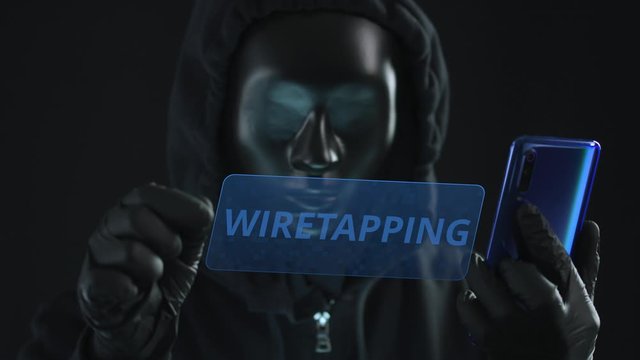 Hacker wearing black mask pulls WIRETAPPING tab from a smartphone. Hacking concept