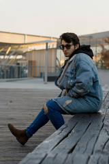 Young fashionable man sitting in near of shopping center in a city. Wears jeans outfit and sunglasses.