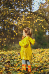 Little funny girl in stylidh yellow raincoat, jeans and rubber boots stands in the autumn forest or park outdoors eating cupcake. Maple leaves foliage on the ground. Concept fall family photo shoot