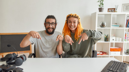 Cheerful man and woman pointing down asking followers to subcribe. Young male and female technology blogger recording video blog or vlog at home