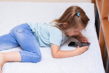 Curious little girl in blue clothes, uses phone applications, looking at a mobile device, lies on the bed.Clear girl plays in a smartphone online game alone at home.Concept of a child and a gadget