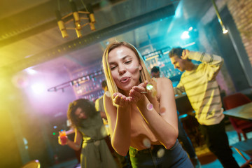 Dance and Party. Attractive caucasian young woman blowing confetti to camera while posing in the night club. Friends dancing, celebrating, having fun in the background