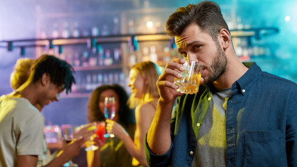 Handsome young man looking at camera while drinking, posing with a cocktail in his hand and friends chatting, having drinks at the bar counter in the background