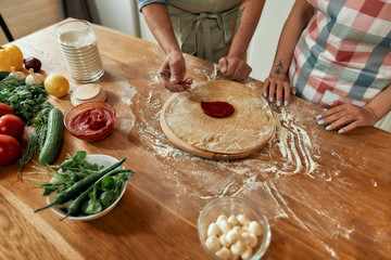Cropped shot of couple making pizza together. Man in apron adding, applying tomato sauce on the dough while woman helping him in the kitchen. Hobby, lifestyle