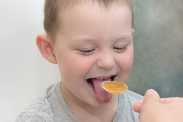 close-up of a child with a sticking out tongue and a funny expression on his face, he is being given medicine on a spoon. The idea is that you need to take medications to stay healthy.