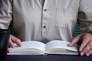 A man in a beige shirt is reading a book at the table. Glasses, watch and cup.