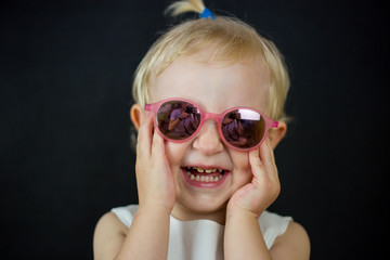 little girl on a black background with glasses on her eyes, a sincere childish smile on her face and blonde hair