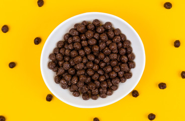 White bowl with chocolate corn cereal balls and milk on yellow background, top-down view. Modern fresh breakfast image.