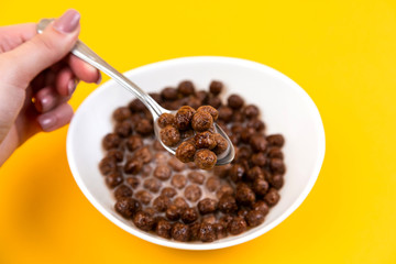 Woman hand holding spoon at White bowl with chocolate corn cereal balls and milk on yellow background, top-down view. Modern fresh breakfast image.