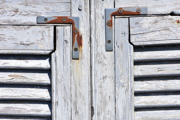 Weathered wooden window shutters with white paint peeling off and rusty strengthening metal pieces.
