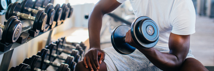Young African American man sitting and lifting a dumbbell close to the rack at gym. Male weight...