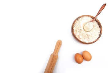 Baking ingredients: Wheat flour in a bowl or wooden plate, brown eggs and a rolling pin on a white background. The view from the top.