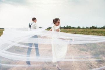 Happy bride and groom run through a wild field with a transparent white veil
