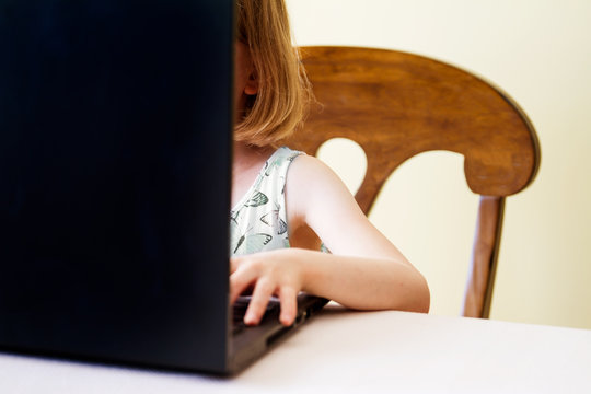 Small girl works on her school at home on a laptop; Child sits at a table with laptop computer and types