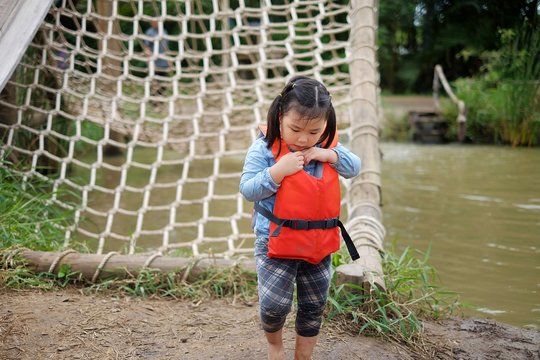 A cute young Asian girl is adjusting her life jacket before climbing up a net over water, having fun but staying safe.