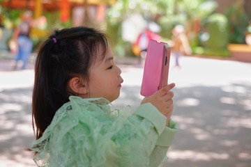 A cute young Asian girl is holding a pink smartphone, trying to take a picture on her vacation.