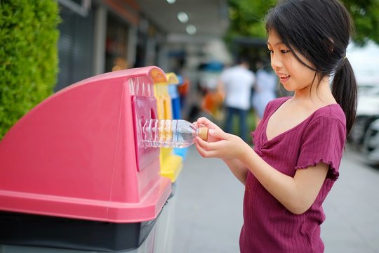 A close up picture of a cute young Asian girl throwing away an empty plastic water bottle on a recycling bin which sorted waste into 3 groups, recyclable bottles and cans, paper and trash.