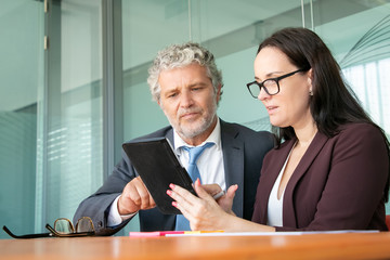 Serious focused colleagues using tablet together, looking and pointing at gadget screen while sitting at table in office. Communication or teamwork concept