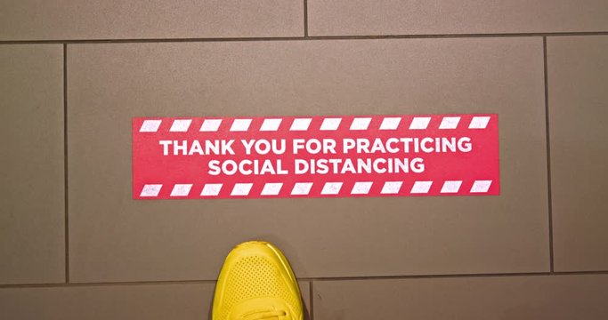Tourist steps on red floor sign at hotel reminding to practicing social distancing. Man in bright sport shoes is walking in hotel lobby by COVID-19 prevention signage. Safe travels during coronavirus