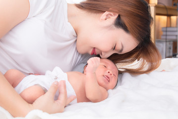 Beautiful happy Asian women mom kissing healthy toddler newborn baby in bed at home, Healthcare medical lifestyle mother's day/ young motherhood concept, infant sleep  safe and protection in mom's arm