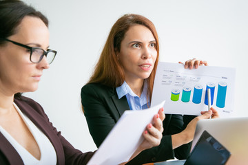 Serious businesswomen analyzing diagrams in office, presenting paper report at meeting, pointing...