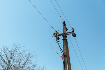  Power line post with electricity cables against a clear sky with white clouds. A tree grows near a pillar. Electricity transmission line, power supply