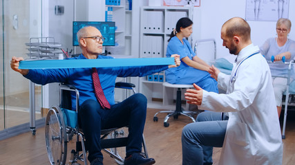 Handicapped injured man training with elastic band under strict medical supervision in modern private recovery facility. Invalid physiotherapy program, health care injury rehabilitation