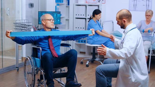 Handicapped Injured Man Training With Elastic Band Under Strict Medical Supervision In Modern Private Recovery Facility. Invalid Physiotherapy Program, Health Care Injury Rehabilitation