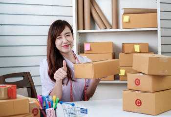 Starting up small business SME, a beautiful young Asian entrepreneur poses a thumbs up to confirm the quality of her merchandise, with a cartons placed on the desk. Work from home concept