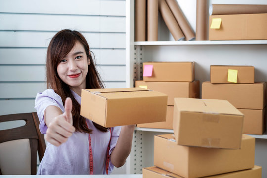 Starting Up Small Business SME, A Beautiful Young Asian Entrepreneur Poses A Thumbs Up To Confirm The Quality Of Her Merchandise, With A Cartons Placed On The Desk. Work From Home Concept