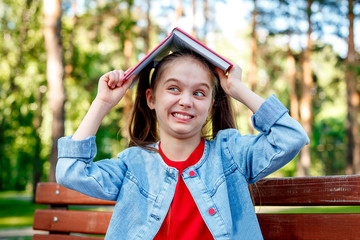 Child Girl With Book Over Her Head Shows a Funny Face Outdoors