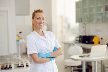 Attractive blond smiling female lab assistant standing in laboratory with hands crossed and looking at camera.