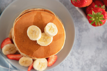 Close-up Top view of a mountain of fresh homemade pancakes, garnished with strawberries and banan, sprinkled with syrup, on a light background. Traditional American, rustic breakfast.