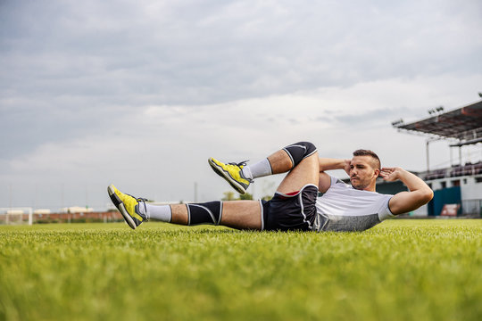 Handsome unshaven soccer player in shape doing sit-ups on the field. - Powered by Adobe