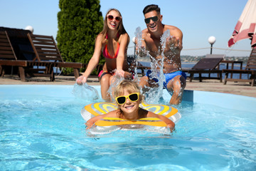 Little boy swimming with inflatable ring near his parents in outdoor pool on sunny summer day
