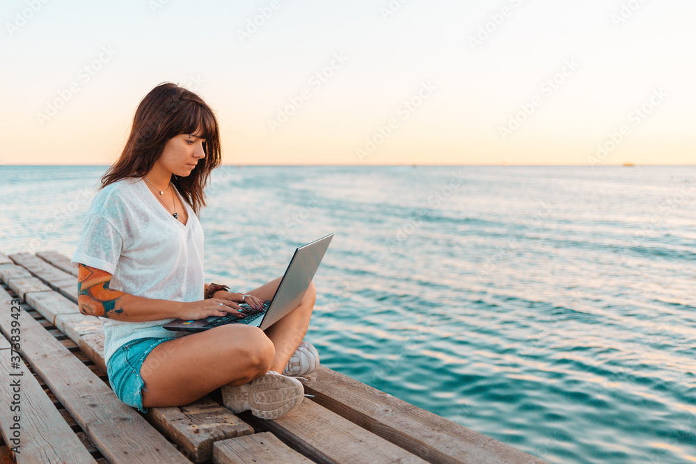 Wall mural a young beautiful woman with tattoos is sitting on a pier by the sea and working at a laptop. sea an