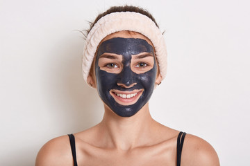 Beautiful smiling woman with black clay facial mask on face, looking at camera with happy expression, having hairband on her head, posing isolated over white background.