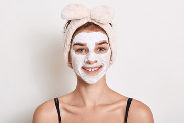 Young woman putting face cream while posing isolated over white background, lady dresses hairband with bow, looking smiling at camera, doing skin care procedures.