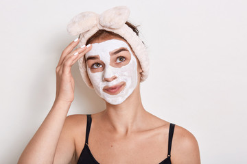 Woman with cosmetics mask applied looking up and touching her eyebrow, having pensive facial expression, doing beauty procedures at home, stands against white background.