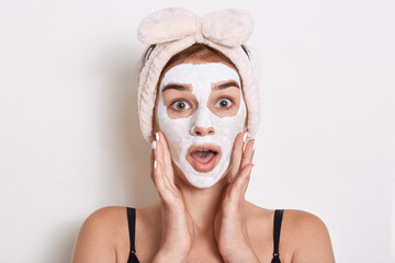 Pretty girl with facial mask, surprised female with headband with bow, lady having astonished expression, posing isolated over white background. Skin care and beauty.